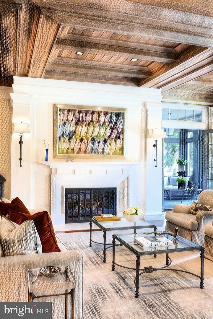 Living room with a rough-hewn beamed wood ceiling, a carved stone fireplace, and a wall of windows to the grounds